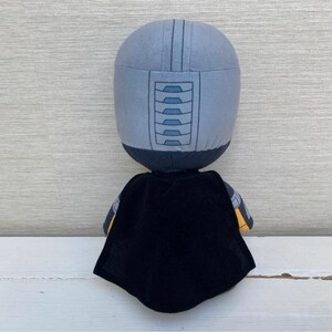 May include: A plush doll with a gray helmet-like head and a black cape. The head has a series of rectangular shapes on the front. The doll's arms have yellow and gray accents. The doll is standing on a white surface.