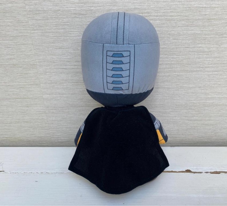 May include: A plush doll with a gray helmet-like head and a black cape. The head has a series of rectangular shapes on the front. The doll's arms have yellow and gray accents. The doll is standing on a white surface.