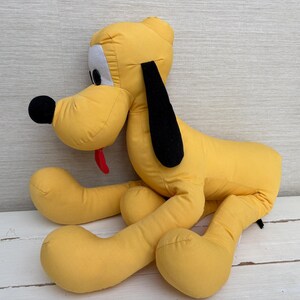 May include: A yellow plush dog toy with long black ears, a black nose, and a red tongue. The stuffed animal features white patches around the eyes and is constructed from fabric, depicted in a seated position with extended front and back legs.