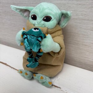 May include: A plush toy of a character with large ears and big eyes, holding a teal-colored creature. The character is light green with a tan robe. The creature has black stripes and orange accents.