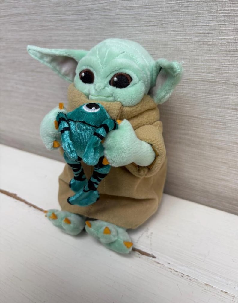 May include: A plush toy of a character with large ears and big eyes, holding a teal-colored creature. The character is light green with a tan robe. The creature has black stripes and orange accents.