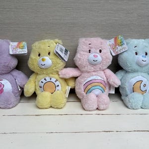 May include: Four plush Care Bears in pastel colors: purple, yellow, pink, and blue. Each bear has a unique belly badge and a tag. The purple bear has a milkshake, the yellow bear has a sun, the pink bear has a rainbow, and the blue bear has a moon.