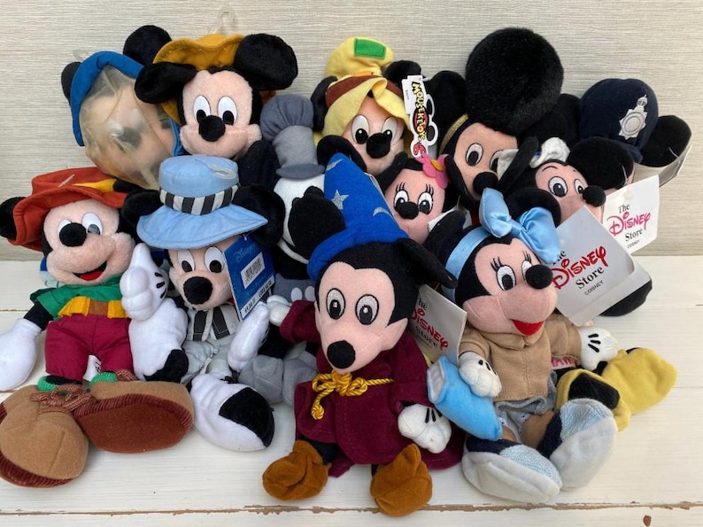 May include: A selection of Mickey Mouse and Minnie Mouse soft toys in different costumes. The plush toys include Mickey Mouse dressed as a sorcerer, a conductor, and other characters. The toys are in various colours, including red, blue, and yellow.