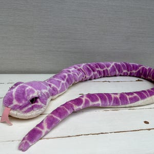 May include: A plush snake toy featuring a vibrant purple and white diamond-patterned fabric. The stuffed animal has a soft, textured exterior, a light-coloured underbelly, dark eyes, and a protruding pink felt tongue extending from its mouth.