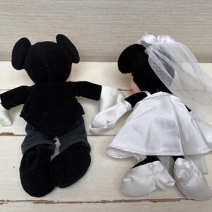 May include: Two plush Mickey and Minnie Mouse dolls. Mickey is dressed in a black tuxedo with white gloves. Minnie is in a white satin wedding dress with a veil. The dolls are positioned as if holding hands, showcasing a wedding theme.