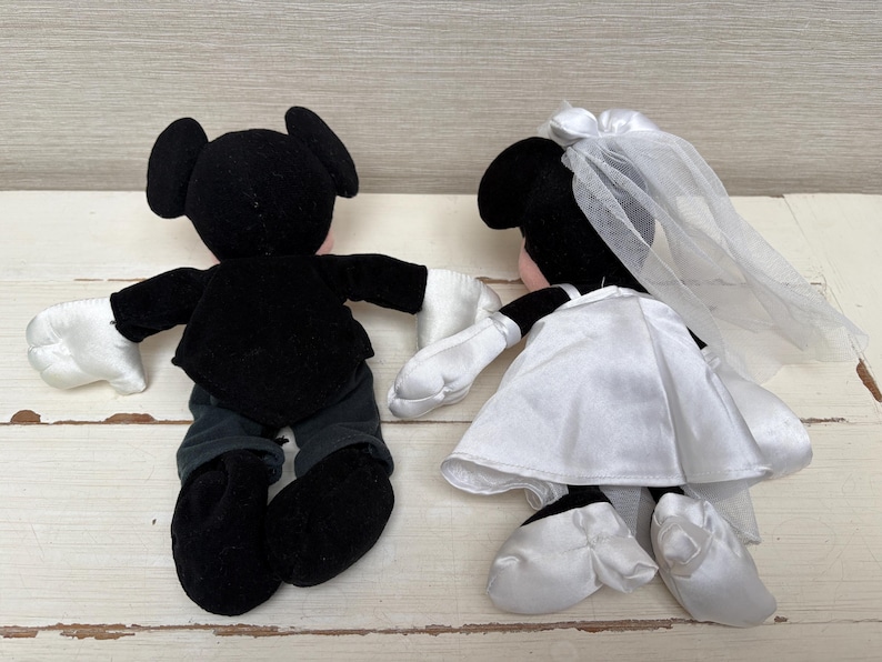 May include: Two plush Mickey and Minnie Mouse dolls. Mickey is dressed in a black tuxedo with white gloves. Minnie is in a white satin wedding dress with a veil. The dolls are positioned as if holding hands, showcasing a wedding theme.
