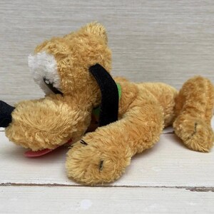 Pode incluir: Um brinquedo de pelúcia de um cão, com corpo castanho, orelhas e nariz pretos. O cão tem uma mancha branca na cara e uma língua rosa. O brinquedo está deitado numa superfície branca.
