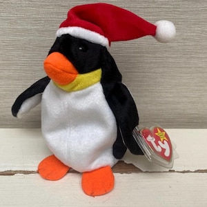 May include: A soft penguin toy wearing a red Santa hat, with black wings and orange feet and beak. The penguin has a white belly and a yellow collar. A clear tag with a red heart and yellow star is attached.