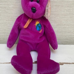 Puede incluir: Un oso de peluche morado con nariz y ojos negros. El oso tiene un diseño bordado de un sol y un globo en el pecho, con el número "2000" debajo. Tiene una cinta dorada alrededor del cuello y una etiqueta sujeta a la oreja.