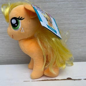 Könnte beinhalten: Ein Plüschpferd in Orange und Gelb mit langer, fließender Mähne und Schweif. Das Pony hat große Augen mit grünen Iris und schwarzen Wimpern. Ein Etikett ist am Spielzeug befestigt.