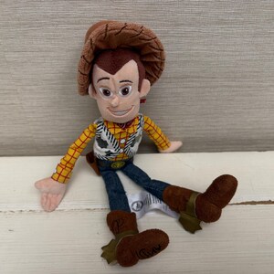 May include: A plush toy of the character Woody from the movie Toy Story. Woody is wearing a brown cowboy hat, a yellow plaid shirt, a white vest with black cow print, blue jeans, and brown boots. The toy is sitting on a white surface.