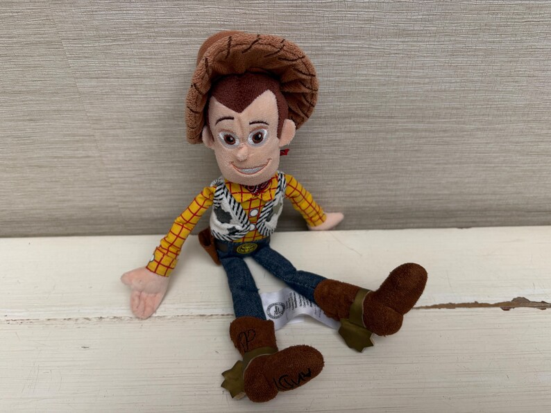 May include: A plush toy of the character Woody from the movie Toy Story. Woody is wearing a brown cowboy hat, a yellow plaid shirt, a white vest with black cow print, blue jeans, and brown boots. The toy is sitting on a white surface.