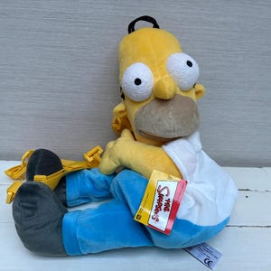 May include: A plush Homer Simpson doll in a seated position. The doll has a yellow head, large white eyes, and a brown nose and mouth. He wears a white shirt, blue trousers, and grey shoes. A yellow backpack is attached.