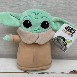 Peut inclure: Peluche Grogu, &eacute;galement connu sous le nom de Baby Yoda, de Star Wars: The Mandalorian. La peluche est vert clair avec de grands yeux noirs, des v&ecirc;tements beiges et de grandes oreilles. Une &eacute;tiquette Star Wars est attach&eacute;e.