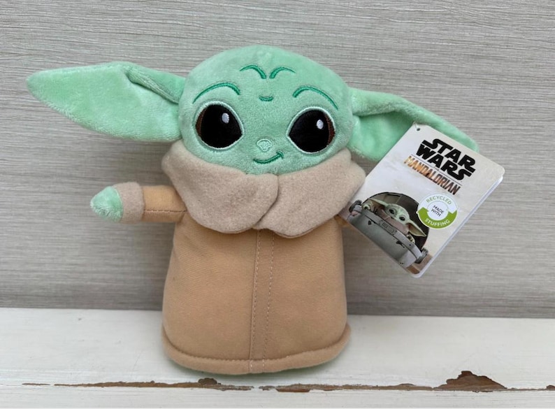 Peut inclure: Peluche Grogu, &eacute;galement connu sous le nom de Baby Yoda, de Star Wars: The Mandalorian. La peluche est vert clair avec de grands yeux noirs, des v&ecirc;tements beiges et de grandes oreilles. Une &eacute;tiquette Star Wars est attach&eacute;e.