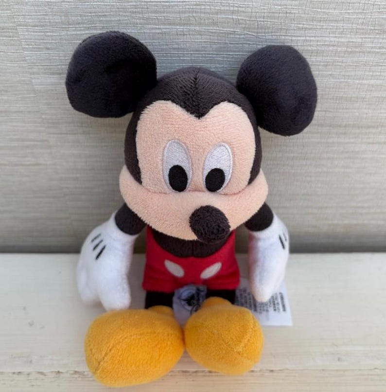 K&ouml;nnte beinhalten: Ein Pl&uuml;schtier von Micky Maus. Das Spielzeug hat schwarze Ohren, einen schwarzen Kopf und ein hellbraunes Gesicht. Es tr&auml;gt ein rotes Hemd, wei&szlig;e Handschuhe und gelbe Schuhe. Das Spielzeug ist etwa 15 cm gro&szlig;.