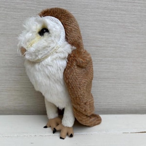 Könnte beinhalten: Eine Plüsch-Eulenfigur mit weißem Gesicht und Brust, braunen Flügeln und Rücken sowie schwarzen Krallen. Die Eule hat ein detailliertes, strukturiertes Aussehen mit einem kleinen Schnabel und dunklen Augen. Die Eule steht auf einer weißen Oberfläche.