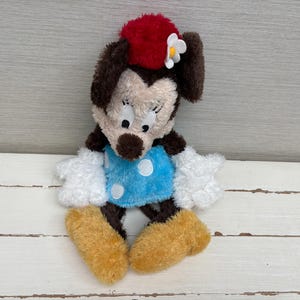 May include: A plush Minnie Mouse doll with a red bow, white flower, and blue dress with white polka dots. The doll has brown ears, a tan face, white gloves, and tan shoes. The toy is sitting on a white wooden surface.