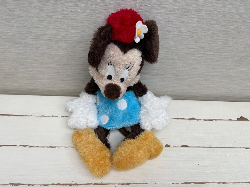 May include: A plush Minnie Mouse doll with a red bow, white flower, and blue dress with white polka dots. The doll has brown ears, a tan face, white gloves, and tan shoes. The toy is sitting on a white wooden surface.