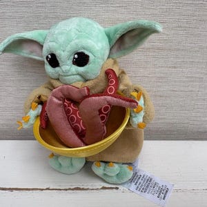 Pode incluir: Uma boneca de pel&uacute;cia Grogu, tamb&eacute;m conhecida como Baby Yoda, com grandes olhos pretos e orelhas grandes. A boneca est&aacute; segurando uma tigela amarela com tent&aacute;culos vermelhos e rosa. A boneca est&aacute; vestindo uma t&uacute;nica marrom e tem pele verde.