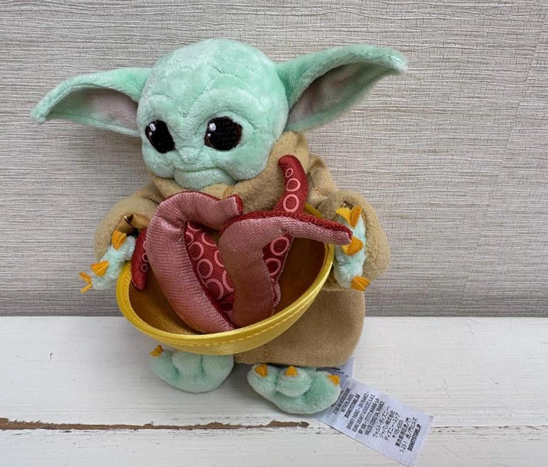 Pode incluir: Uma boneca de pel&uacute;cia Grogu, tamb&eacute;m conhecida como Baby Yoda, com grandes olhos pretos e orelhas grandes. A boneca est&aacute; segurando uma tigela amarela com tent&aacute;culos vermelhos e rosa. A boneca est&aacute; vestindo uma t&uacute;nica marrom e tem pele verde.