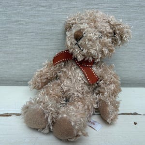 May include: A small, plush teddy bear with light brown fur and a dark red ribbon tied around its neck. The bear has a stitched nose and a tag attached to its foot. The toy is sitting on a white surface.