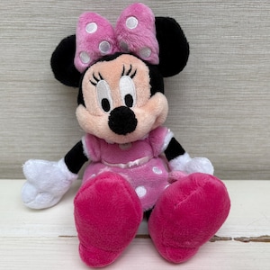 May include: A plush Minnie Mouse doll, featuring a pink dress with white polka dots, a matching bow with white polka dots, and large pink feet. The doll has black ears, arms, and a black nose. The eyes are white with black eyelashes.