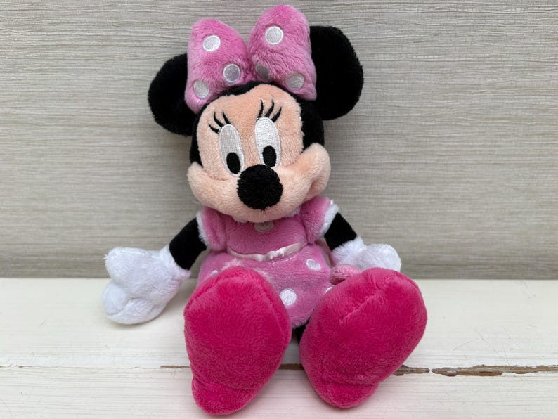 May include: A plush Minnie Mouse doll, featuring a pink dress with white polka dots, a matching bow with white polka dots, and large pink feet. The doll has black ears, arms, and a black nose. The eyes are white with black eyelashes.