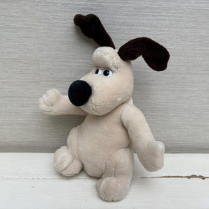Könnte beinhalten: Ein Plüschhund mit cremefarbenem Körper, zwei dunkelbraunen Schlappohren und einer großen, runden, schwarzen Nase. Die Figur hat weiße Augen mit kleinen schwarzen Pupillen, eine weiche Stoffoberfläche und befindet sich in sitzender Position.