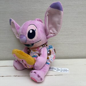 Puede incluir: Mu&ntilde;eco de peluche de Angel, personaje de Lilo & Stitch, de color lavanda. Tiene grandes orejas y sostiene un donut amarillo. Viste un vestido con estampado de donuts. Un juguete adorable y reconocible.