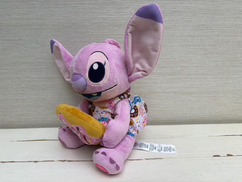 Puede incluir: Mu&ntilde;eco de peluche de Angel, personaje de Lilo & Stitch, de color lavanda. Tiene grandes orejas y sostiene un donut amarillo. Viste un vestido con estampado de donuts. Un juguete adorable y reconocible.