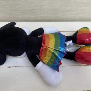 May include: A black plush character with a red bow, white gloves, and a rainbow-striped skirt. The character's shoes are red with rainbow stripes. The toy is on a white wooden surface.
