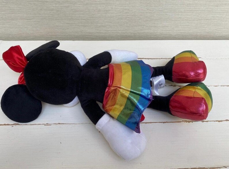 May include: A black plush character with a red bow, white gloves, and a rainbow-striped skirt. The character's shoes are red with rainbow stripes. The toy is on a white wooden surface.