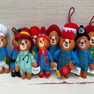 Puede incluir: Una colección de adornos de peluche de Paddington Bear, cada uno vestido con un atuendo único. Los osos llevan varios sombreros, incluyendo un gorro de chef, un uniforme de médico y un casco de bombero. Llevan chaquetas azules y tienen pequeñas etiquetas.