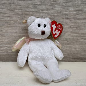 Puede incluir: Un oso de peluche blanco con ojos negros y nariz marrón. El oso tiene alas iridiscentes y una cinta rosa alrededor del cuello. Una etiqueta roja en forma de corazón está sujeta a la oreja del oso.