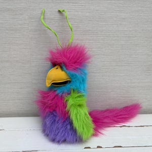 May include: A plush bird puppet featuring vibrant multi-colored faux fur in shades of hot pink, bright blue, lime green, and purple. The puppet has a prominent yellow beak, a long pink tail, and two thin lime green antennae extending from the top.