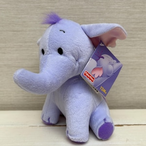 May include: A light purple plush elephant toy with a small tuft of purple hair on its head, large ears with pink inner lining, and dark embroidered eyes. A Fisher-Price brand tag is attached to the ear, featuring an illustration and 12M+ text.