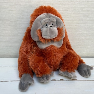 May include: A plush orangutan toy featuring long, shaggy, rust-colored fur and a smooth, light gray face. The stuffed animal has gray hands and feet, a rounded head, and a soft, textured appearance, designed with a seated posture and realistic facial features.