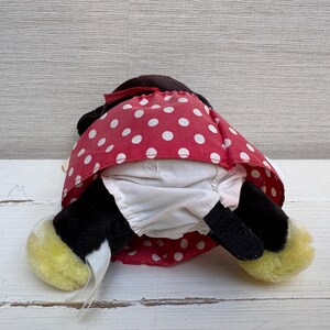 K&ouml;nnte beinhalten: Ein Pl&uuml;schtier einer Zeichentrickfigur, von hinten gesehen. Das Spielzeug hat schwarzes Fell, einen roten Polka-Dot-Rock und gelbe F&uuml;&szlig;e. Das Spielzeug liegt auf einer wei&szlig;en Holzoberfl&auml;che.