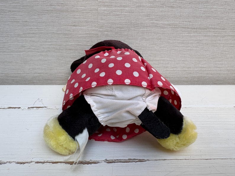 K&ouml;nnte beinhalten: Ein Pl&uuml;schtier einer Zeichentrickfigur, von hinten gesehen. Das Spielzeug hat schwarzes Fell, einen roten Polka-Dot-Rock und gelbe F&uuml;&szlig;e. Das Spielzeug liegt auf einer wei&szlig;en Holzoberfl&auml;che.