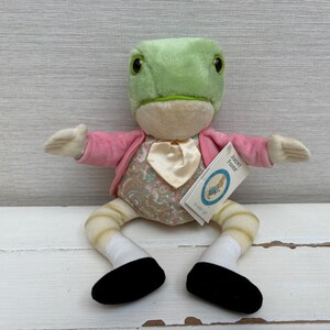 Könnte beinhalten: Ein Frosch-Stofftier mit hellgrünem Kopf, rosa Jacke und gemusterter Weste mit cremefarbener Krawatte. Der Frosch hat gestreifte Beine, weiße Socken und schwarze Schuhe. Ein weißes Etikett an der Jacke trägt die Aufschrift Jeremy Fisher.