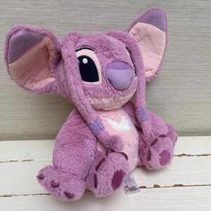 May include: A plush toy of the character Angel from the Disney movie Lilo & Stitch. The toy is primarily pink and purple, with large ears, a distinctive nose, and large black eyes. It has a soft, fuzzy texture and is designed to resemble the animated character.