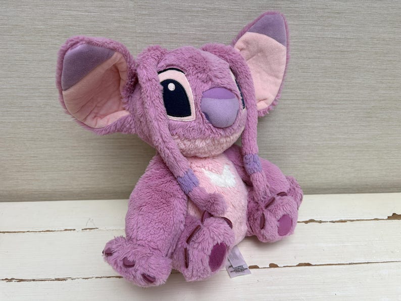 May include: A plush toy of the character Angel from the Disney movie Lilo & Stitch. The toy is primarily pink and purple, with large ears, a distinctive nose, and large black eyes. It has a soft, fuzzy texture and is designed to resemble the animated character.