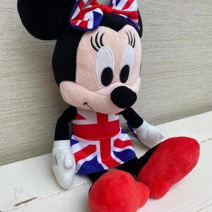 Puede incluir: Un peluche de Minnie Mouse vestida con un atuendo con el tema de la bandera brit&aacute;nica. La mu&ntilde;eca tiene orejas negras y un lazo con un dise&ntilde;o de la bandera brit&aacute;nica. Lleva una falda con el dise&ntilde;o de la bandera brit&aacute;nica y zapatos rojos.