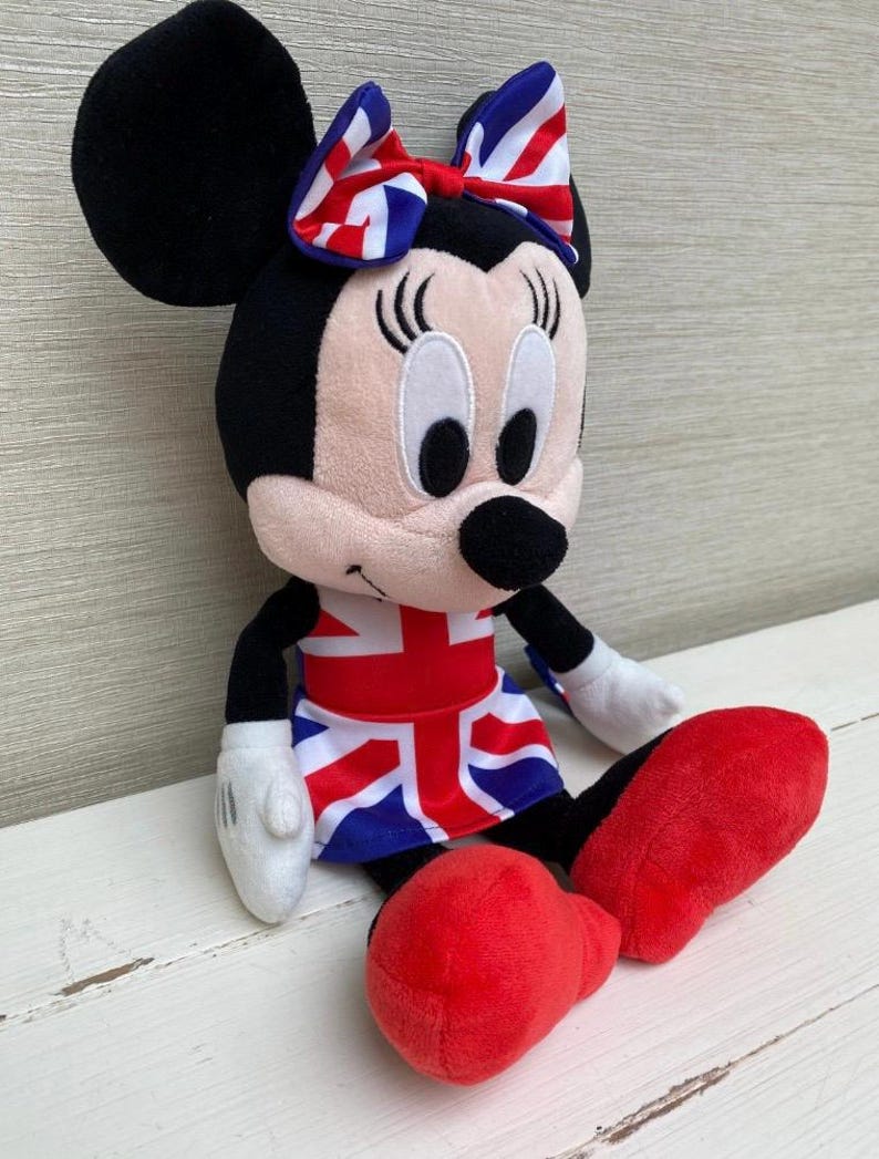 Puede incluir: Un peluche de Minnie Mouse vestida con un atuendo con el tema de la bandera brit&aacute;nica. La mu&ntilde;eca tiene orejas negras y un lazo con un dise&ntilde;o de la bandera brit&aacute;nica. Lleva una falda con el dise&ntilde;o de la bandera brit&aacute;nica y zapatos rojos.