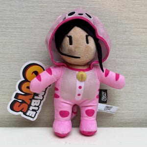 May include: A plush toy of a character wearing a pink cat costume. The toy has black hair and a beige face. The costume features pink stripes and a yellow bell. A tag with the words "Stumble Guys" is attached.