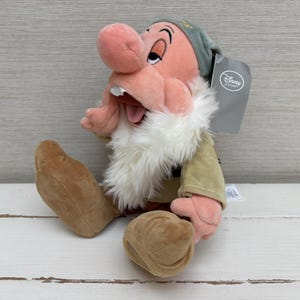 Op de afbeelding: Een knuffel van het personage Dopey uit Sneeuwwitje en de zeven dwergen, met een grote roze neus, een witte baard van imitatiebont, een groene muts en lichtbruine schoenen. Een grijs Disney Store-label is aan de zijkant van het hoofd bevestigd.
