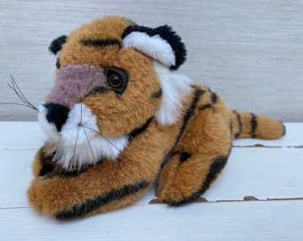 May include: A soft toy tiger cub with brown and black stripes, white details, and a pink nose. The plush animal has a furry texture and realistic features, including whiskers and dark eyes. The toy is lying down on a light-coloured surface.