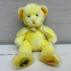 May include: A yellow plush teddy bear with a light yellow ribbon around its neck. The bear has a Scorpio symbol on its foot. The bear is sitting on a white wooden surface.