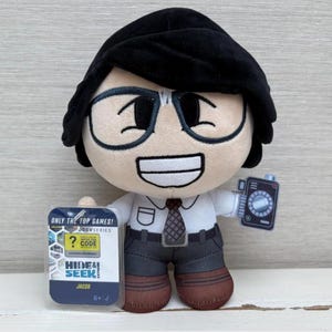 Può includere: Un peluche di un personaggio con occhiali, camicia bianca e cravatta, che tiene in mano una carta di gioco e un piccolo dispositivo. La carta di gioco dice "HIDE & SEEK!" e "JACOB". Il personaggio ha i capelli neri e sorride.